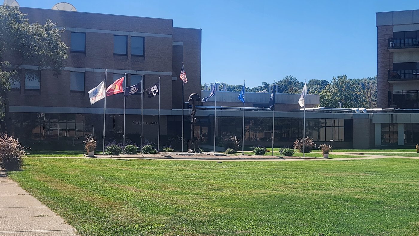 Mitchell Hall at the Indiana Veterans Home with military branch flags and a memorial sculpture on the front lawn